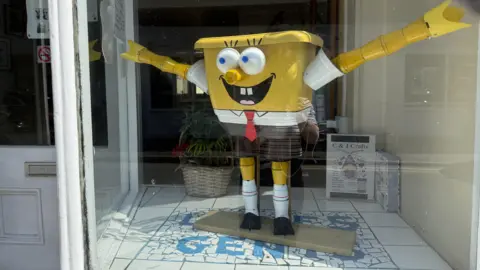 BBC/Amber Gash A square yellow statue wearing a white shirt, red tie and shorts stands with its arms open wide in the window of a shop.