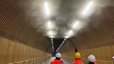 Smoke gathered at the top of a tunnel in Jersey with three people wearing hard hats and orange high vis jackets looking up at it.