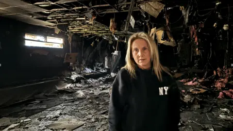 BBC Louise Edge, dressed in a black hoodie, stands in the charred wreckage of her dance school