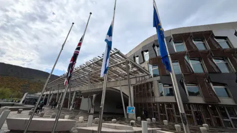 Flags outside the Scottish Parliament flying at half mast. The Union Jack is on the left, the Scottish Saltire in the centre and the European flag on the right. The parliament building is in the background.