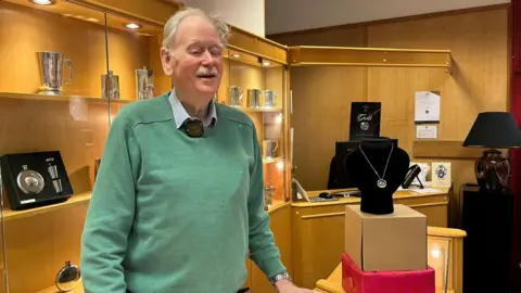 A man in a light green jumper stands in a jewellery shop smiling
