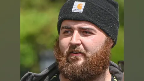 Jamie Fulford. He has a black beanie hat on, and a ginger beard. He is wearing a black coat and looking away from the camera. 
