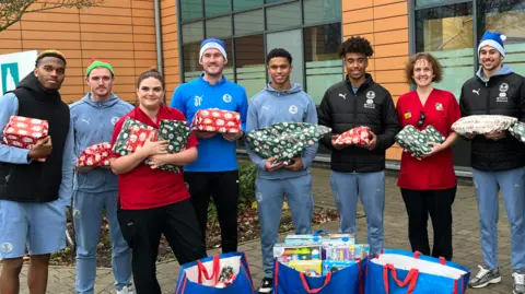 John Devine/BBC Peterborough United players and hospital staff holding presents outside the hospital. 