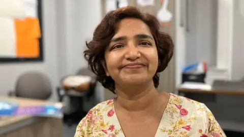 JOANNA TAYLOR/BBC Aparna Nigam is smiling and looking at the camera. She is wearing a floral shirt. There is an out of focus classroom in the background. 