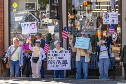 Getty Images Neliela protestētāju grupa ir sapulcējusies pie veikala. Viņiem ir Amerikas karogi un uzraksti, kas lasa "Ja jūs neuztraucaties par mūsu valsti, jūs nepievēršat uzmanību" un "Tā ir valdība, par kuru uztraucās mūsu dibinātāji"