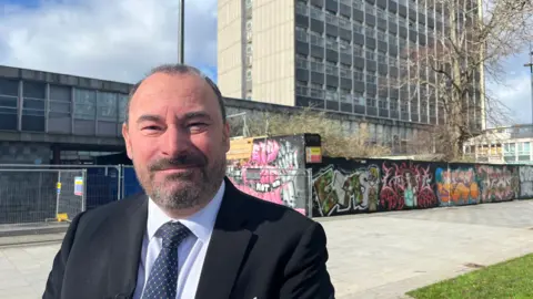 Andy Lugger, also standing near Plymouth Civic Centre. He is a middle-aged man with short, greying hair and a beard. He is wearing a dark suit, a white shirt, and a spotted tie.