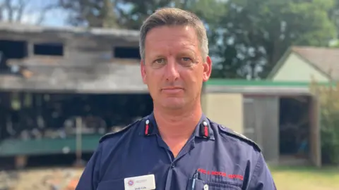 Andy Cole faces the camera. He wears a blue open-necked fire service shirt and has grey hair.