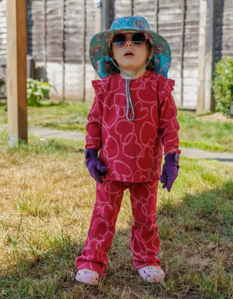 PA Media A child in a red outfit wearing a sun hat, gloves and sunglasses outdoors
