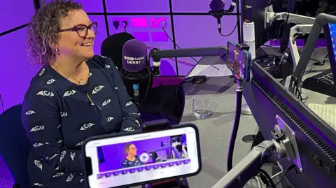 Woman with short curly hair and glasses sat smiling in radio studio with purple microphone infront of her with BBC Radio Derby on it