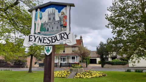 A picture of a sign saying "Eynesbury" on a green