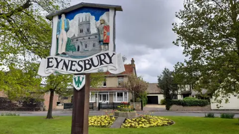 Google A picture of a sign saying "Eynesbury" on a green