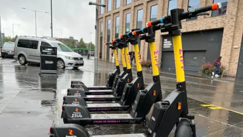BBC A row of yellow scooters with a rainy street and people walking