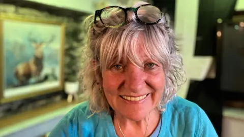 Trina Crawford smiling at the camera. She is wearing a blue t-shirt and has a pair of black glasses on top of her head. In the background is the pub, and a large painting of a deer on the left. 