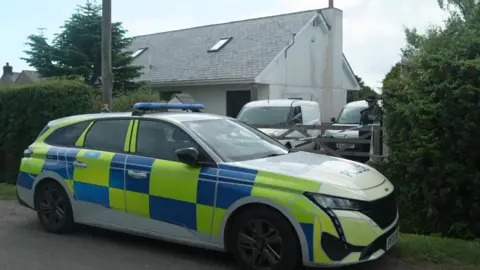 A marked police car is parked outside a rural house, with a gate and hedges partially blocking the driveway. An officer stands near two vans inside the property, suggesting an active police presence at the scene.