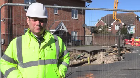 Surrey County Council A man in a high-vis yellow coat with "Surrey County Council" written on the chest with the authority's emblem. He is wearing a white hard hat and standing in front of a metal fence with a large hole the other side.