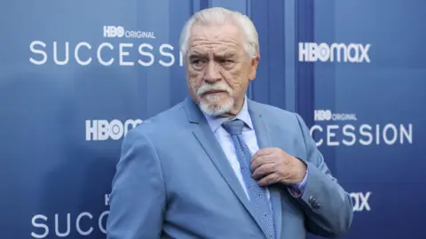 A man with white hair and a white beard looks off to the left. His left hand is across his chest and he is wearing a light blue shirt with a blue tie. 