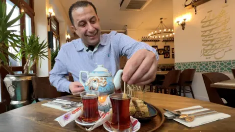 Massood Khamloo serves traditional Iranian tea in his restaurant, Shiraz, on Cowley Road, Oxford. Smiling, he stirs a black tea in a glass with a saffron sugar stick. A plate of baklava is next to the tea. 