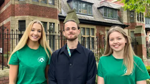 Sofía Luis-Hobbs/BBC Two women and a man smile at the camera in front of a brick building. Both women are stood to either side of the man and both wear matching green tshirts. The man wears a navy overcoat.