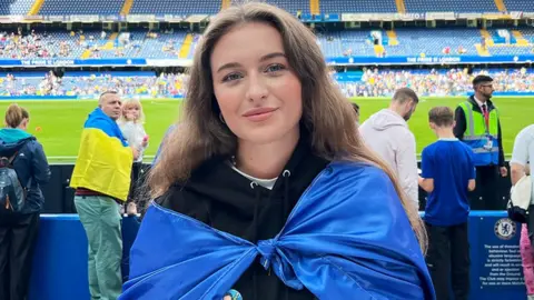 Marta Vasyuta Marta, a Ukrainian woman with a blue and yellow Ukrainian flag around her looking at the camera smiling. She is wearing a black hoody. The background is of Stamford Bridge football stadium, with other people dressed in Ukrainian colours, a green grass pitch and blue seats.