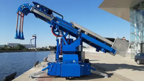 A blue machine on a quayside has a hydraulic arm with electricity cables dangling from the end.