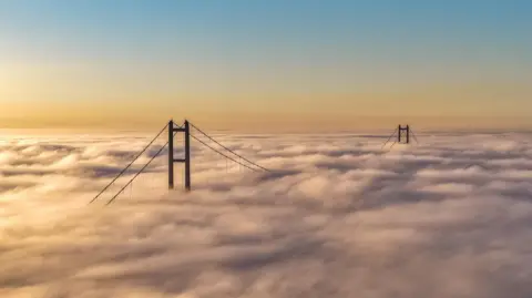 Droneymark The two towers of the Humber Bridge emerge from a blanket of clouds. The sky above is blue fading into orange.