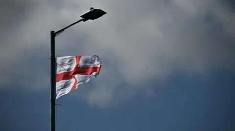 A St George's flag attached to a lampost is blowing in the wind against a slightly grey looking sky. 