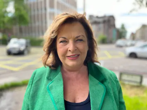 A woman with light brown hair, centred, wearing a green blazer. In a blurred out background, cars can be seen on a road in front of tall buildings. 