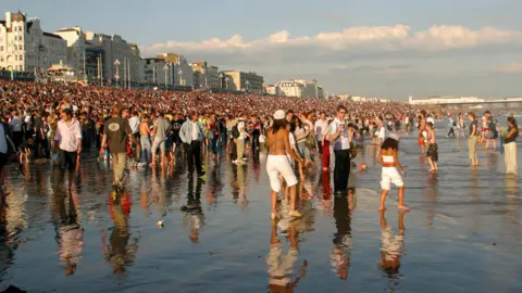 Getty Images A large group of people on a beach. Some people are stood in sea water, which is up to their ankles. A row of houses and a pier are in the background.
