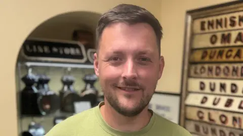 Mark is wearing a green t-shirt. He has brown hair, and brown facial hair. He's standing with train memorabilia in the background.