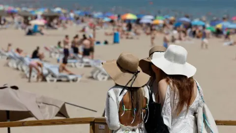 Three women with beach hats stand looking at a beach that has people sunbathing, sat under parasols, and swimming in the sea.