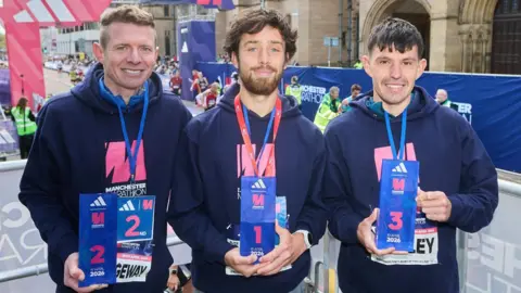Manchester Marathon Left to right - William Strangeway, Yohan Lidove, Charlie Brisley. They're all wearing medals over their navy blue hooded tops and hold blue signs with their respective finishing positions. The finishing arch is behind them next to the University of Manchester stone building.