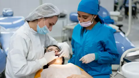 Getty Images Two female dentists carry out an examination on a third woman's mouth. They are wearing blue and white PPE. The patient is lying down with her mouth open.