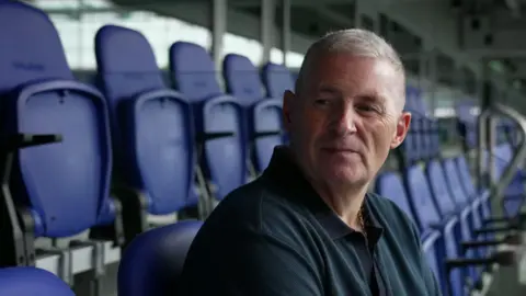 BBC Stephen Simpson, with short grey hair and wearing a navy blue polo shirt, smiles while being interviewed inside Everton's new stadium.