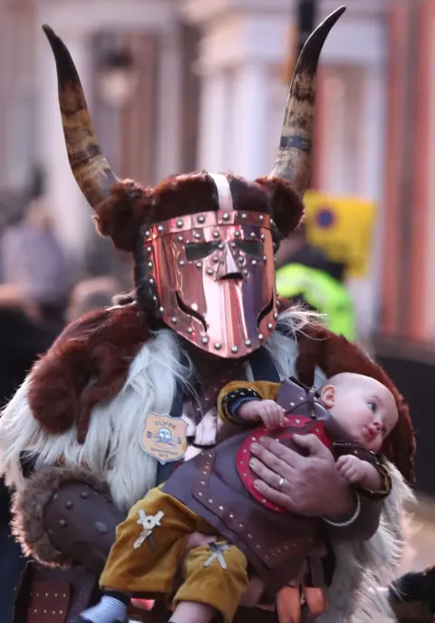 PA Media/Gareth Fuller Reveller dressed as a warrior walks through Lewes carrying a baby