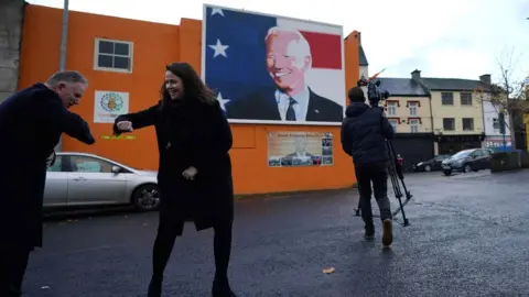 REUTERS/Clodagh Kilcoyne Laurita Blewitt 'elbow bumps' a news crew in Ballina beside a mural of Joe Biden