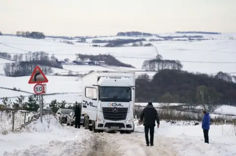 Jacob King/PA Media Motorists gather by a stuck HGV along the snow-covered A515 near Biggin, in the Peak District, Derbyshire, amid freezing conditions in the aftermath of Storm Arwen.