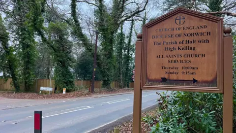 MARTIN GILES/BBC Road surrounded by trees and shrubbery with a wooden sign indicating a church.