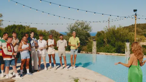 Host Dannii Minogue wears a bright green dress and addresses the cast of the show at a poolside. Fairy lights hang above the group of ten men 