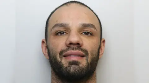 A custody image of Wade Priestley, who has close cropped black hair and a beard