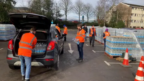 Welsh Water unit   successful  orangish  jackets load   cars with bottled h2o  successful  a supermarket car   parkland  successful  Treorchy.  