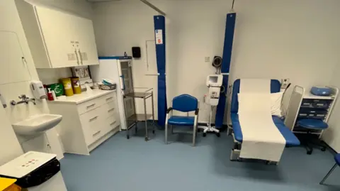 Honiton Surgery An image of a clinical room in Honiton Surgery. It is clean with new storage units, equipment and a sink. There is a blue bed on the right with a blue chair to the left of it. 