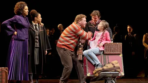 The cast from the play Harry Potter and the Cursed Child on stage. 
There is a girl sitting on a suitcase with a man playing her dad beside her
Behind her is a woman wearing a purple gown standing next to a teenager.
There are a few people standing behind them too.