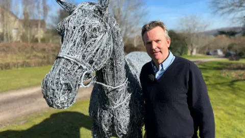 Rupert Till wearing a blue and white checked collared shirt beneath a navy blue jumper. He is standing on a patch of grass alongside a grey steel sculpture depicting a race horse, wearing a bridle and reins. Rupert Till has short grey hair and is smiling at the camera.