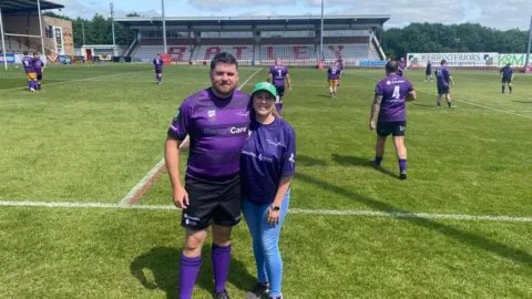 Ben Coombes Ben Coombes is standing on a rugby field with his wife Daisy. Ben is a wearing a purple rugby outfit with other people players behind him in the same uniform. 
His wife daisy is wearing a purple t-shirt, blue jeans and a green cap. 