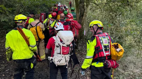 Andrew Vaughan A group of people in hi-vis jackets and helmets standing in a wooded area. Some carry large backpacks. 