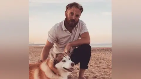 Benjamin Gladman kneeling on a beach with a dog. Mr Gladman, who has light brown hair and a beard, is wearing a beige short-sleeved shirt and blue trousers. He is barefoot. He has his right hand on the back of the dog, which is a German Shepherd-type breed.