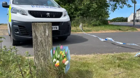 Flowers that lean against a post on the Irvine Beach. A police car is parked right behind.