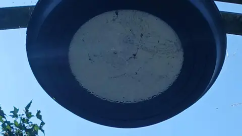Close-up image taken from below of a dark circular-shaped street light. Its surface is scratched and marked. It appears to be in the centre of an arch, although only the immediate supporting structure of two metal bars, extending either side of the lamp, can be seen.
