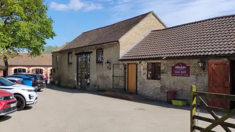 A bricked barn has a car park outside the front on the left. There is a fence on the right which opens into a court yard and there are signs on the door.