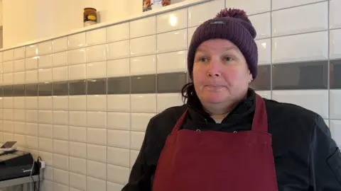 Fishmonger Angela Preece stands in her shop in front of a wall of white tiles, wearing a red apron, black fleece and purple hat. Weighing scales are visible in the background to the left